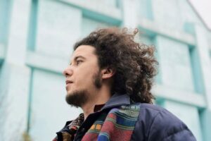 Picture of Mo Kolours wearing a winter coat and a scarf. A modern building is displayed as background